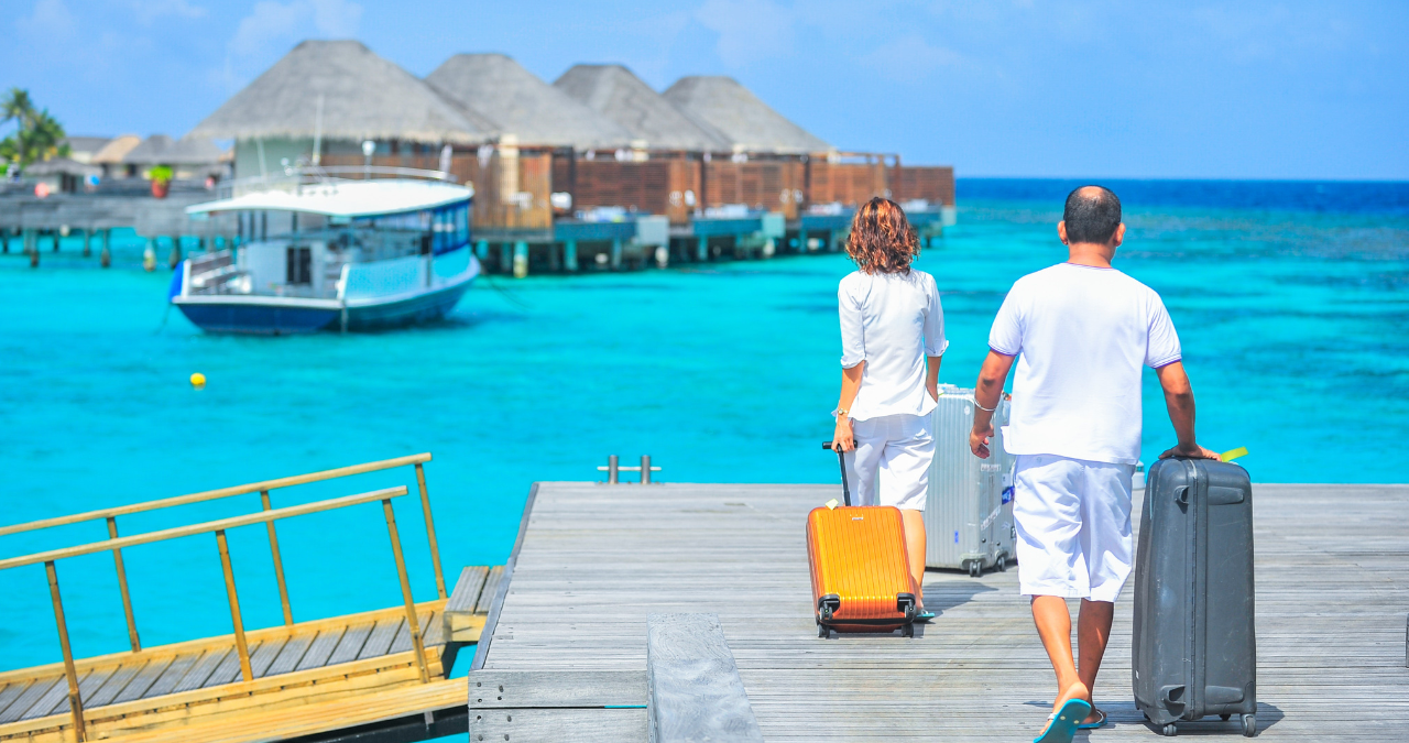 Couple dressed in white with luggage walking towards island resort